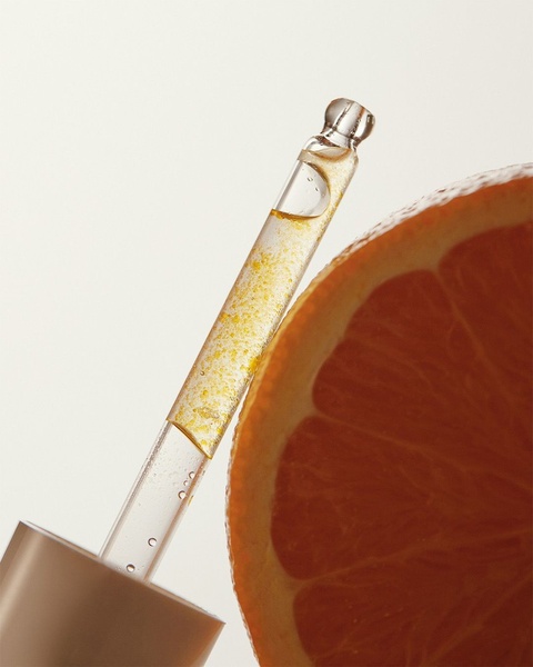 A close-up studio shot featuring a glass dropper filled with a gold-flecked serum next to a fresh orange slice, emphasizing natural ingredients.